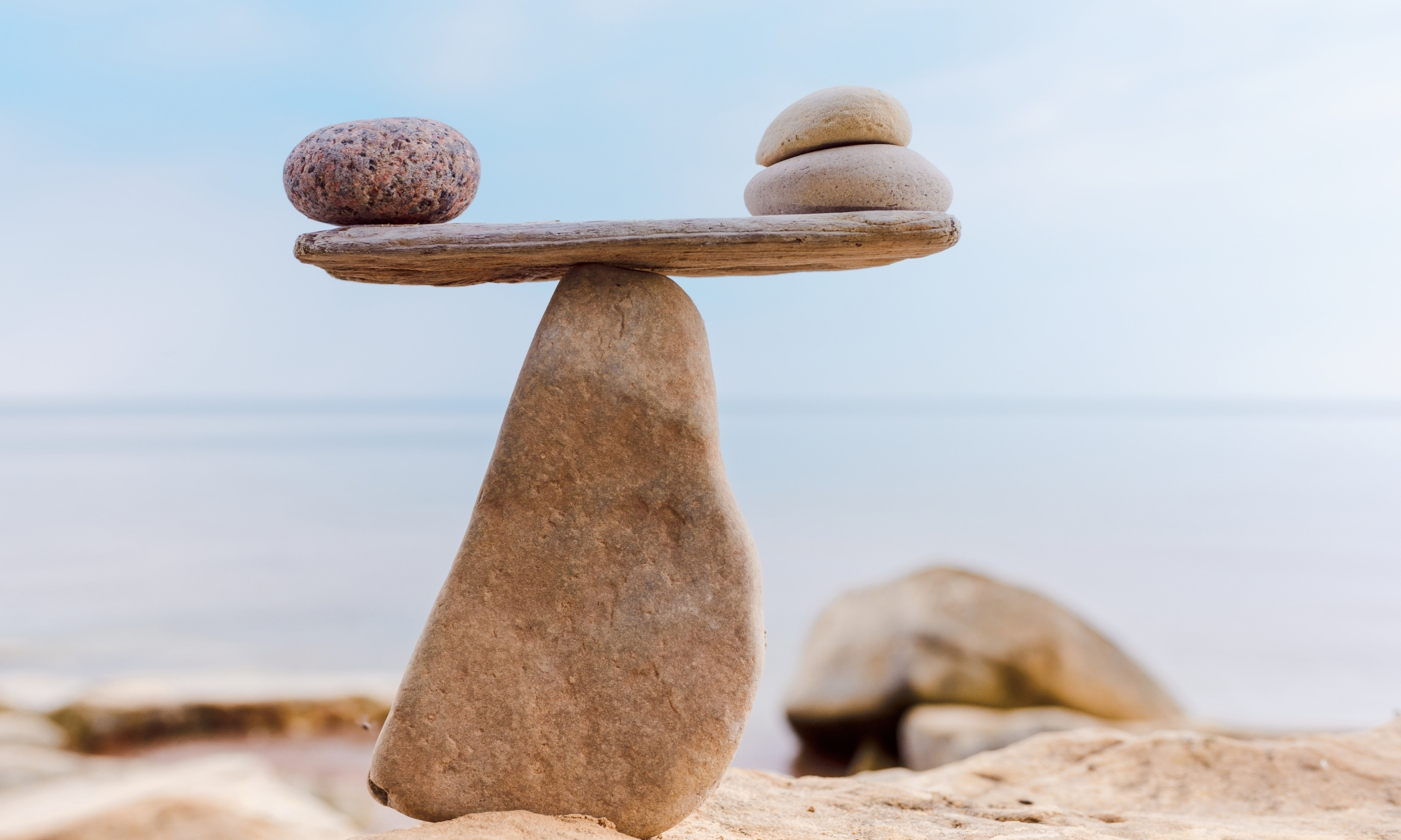 A balanced arrangement of three smooth stones atop a flat rock, forming a scale-like structure by the sea.
