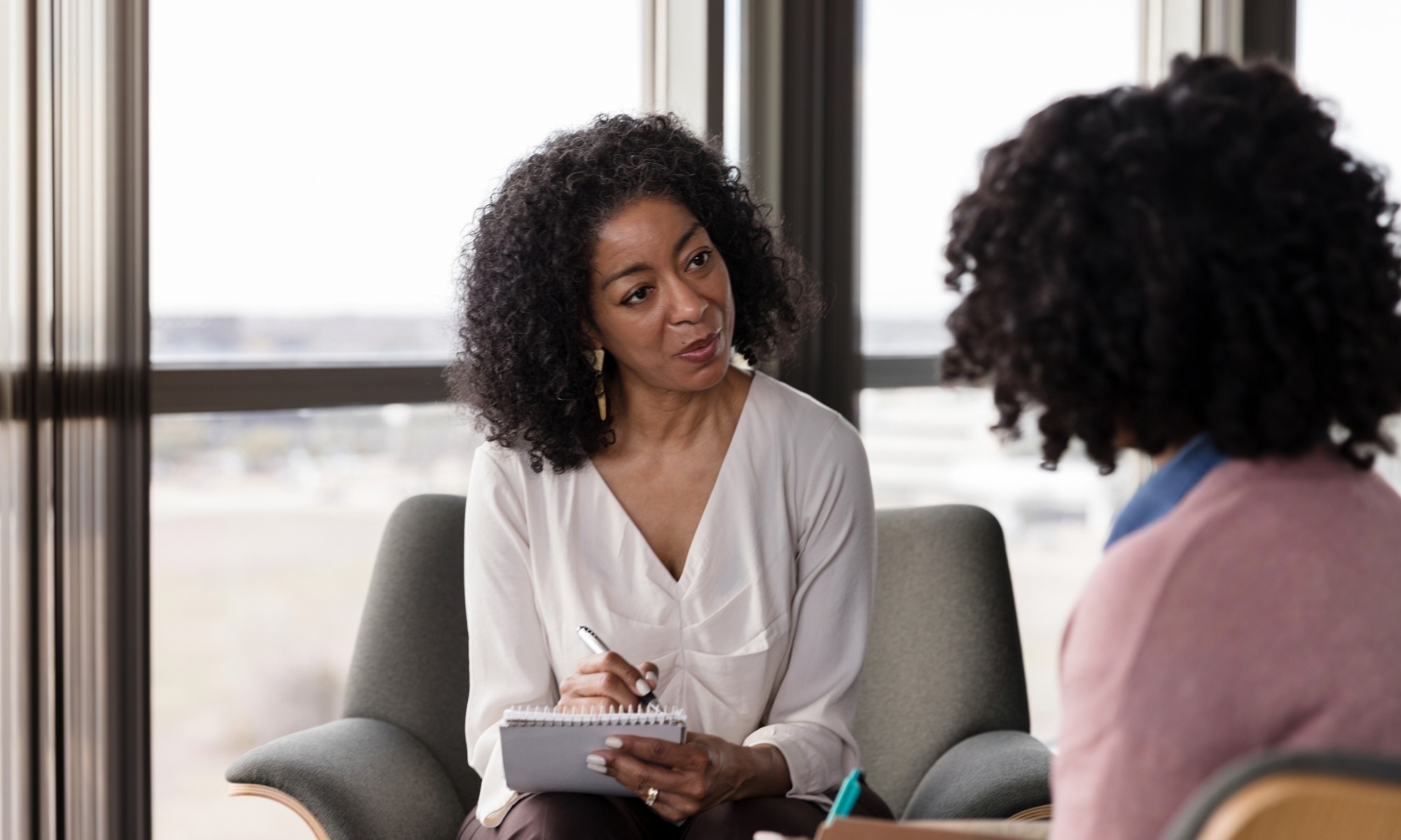 A woman holding a notebook listens attentively to another woman during a conversation in an office setting with large windows in the background.